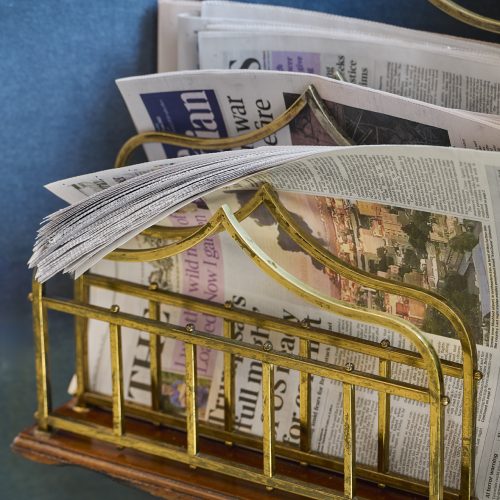 Golden brass newspaper rack holder with magazines and newspapers against blue wall