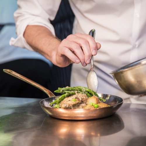 Chef plating gourmet fish dish with sauce and green vegetables in copper pan at Wild Honey Michelin restaurant professional kitchen